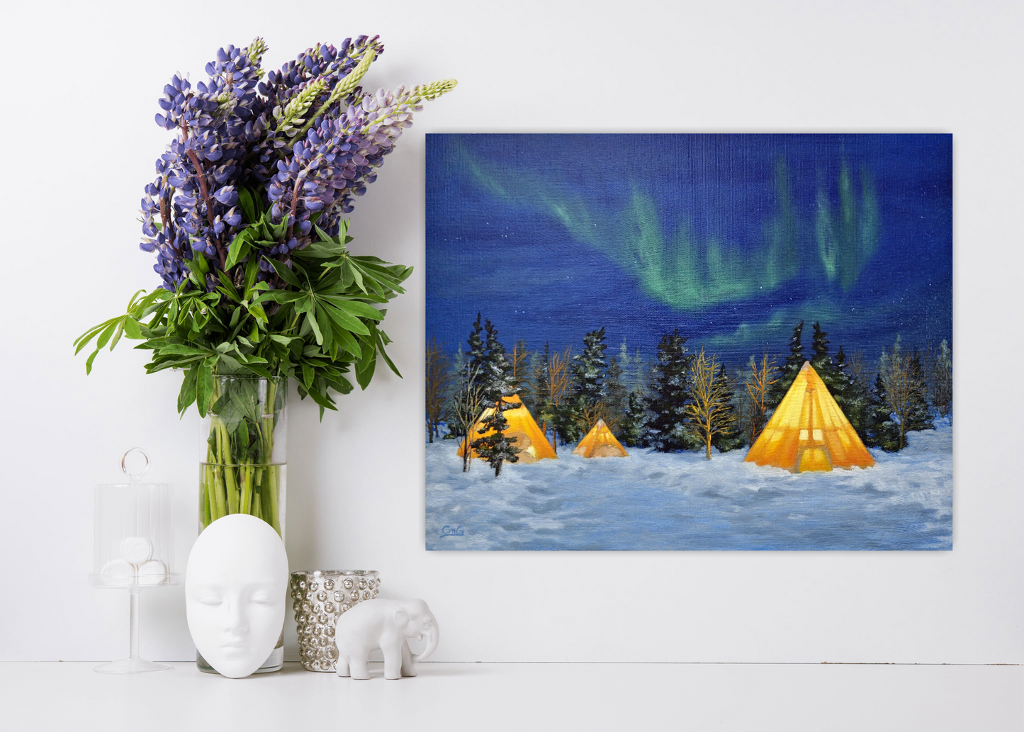 White desk and white wall, with purple flowers in a vase and some nick-nacks and a landscape painting of tipis light with the aurora behind them