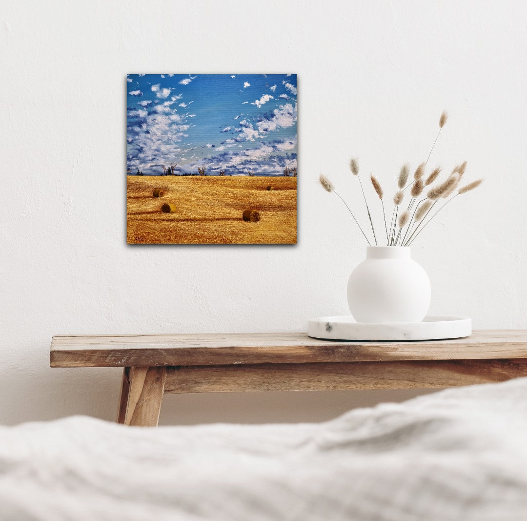 a painting of 4 bales scattered around a golden fall field, with big clouds coming over the horizon, over a wooden bench