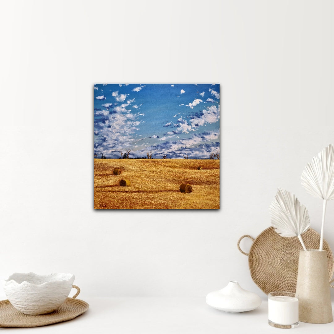 a painting of 4 bales scattered around a golden fall field, with big clouds coming over the horizon over a collection of white pottery and wicker decorations