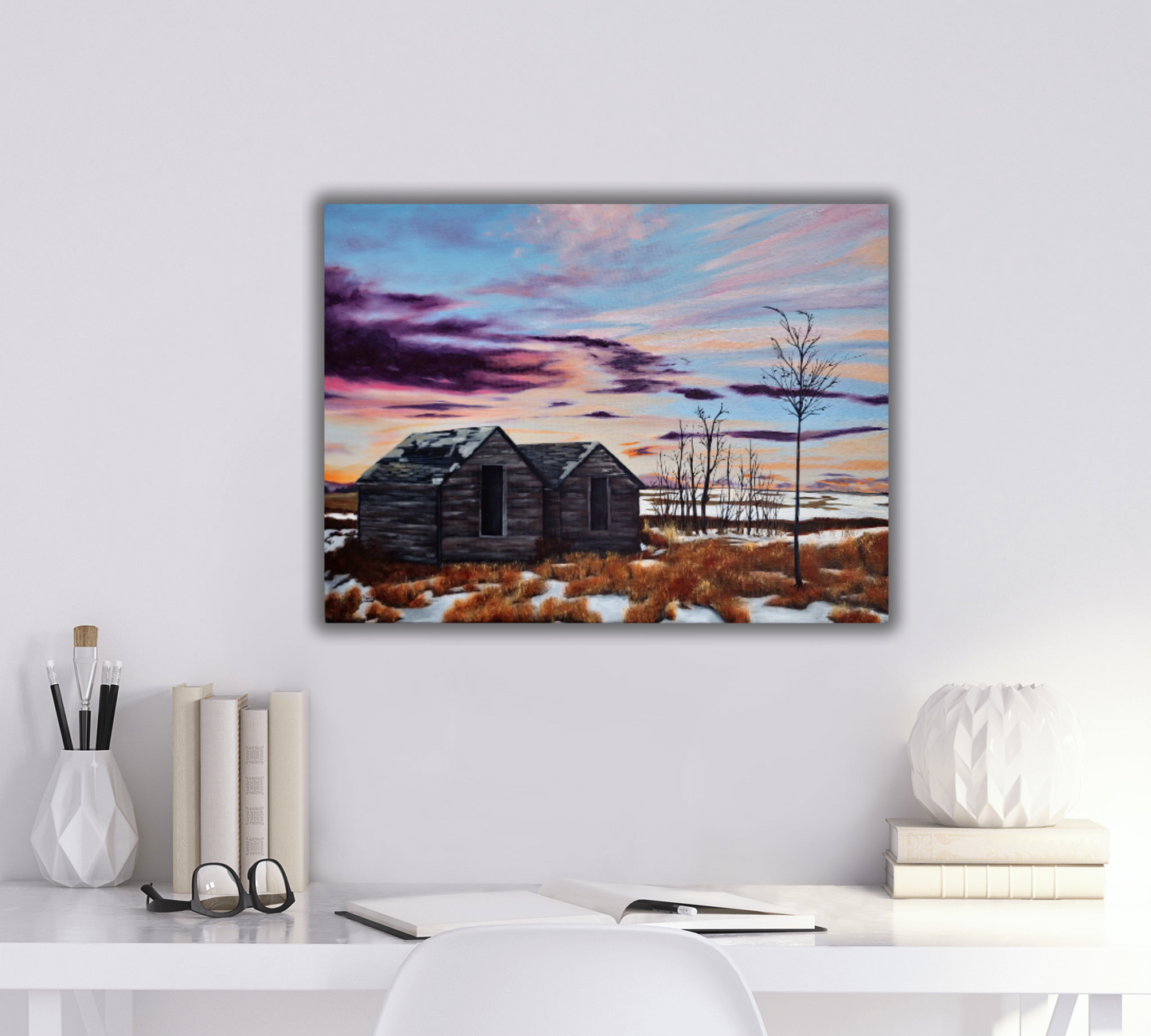 White desk with stationary and glasses on it, and a landscape painting of a  purple and orange sunset with old sheds on the wall