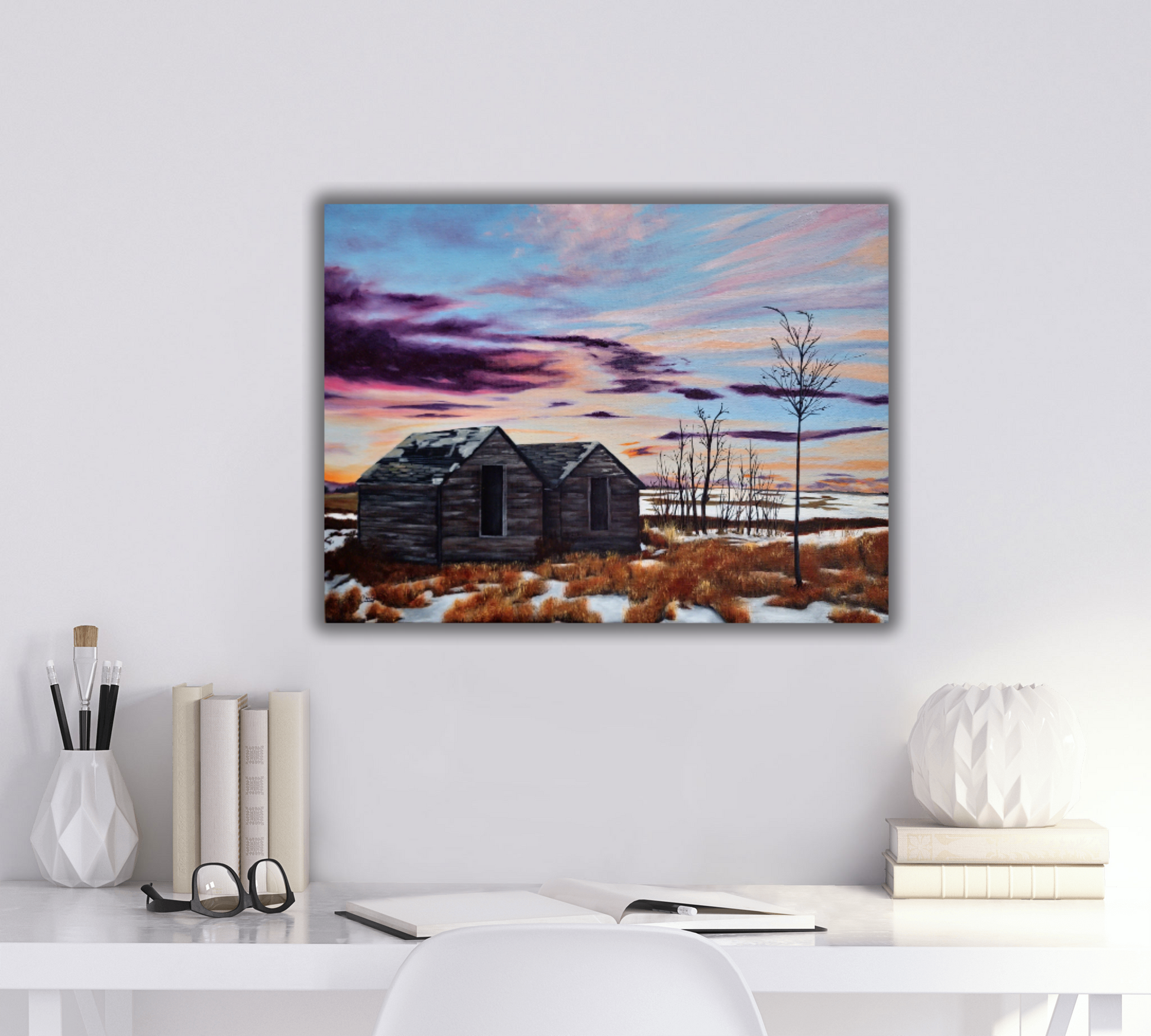 White desk with stationary and glasses on it, and a landscape painting of a  purple and orange sunset with old sheds on the wall