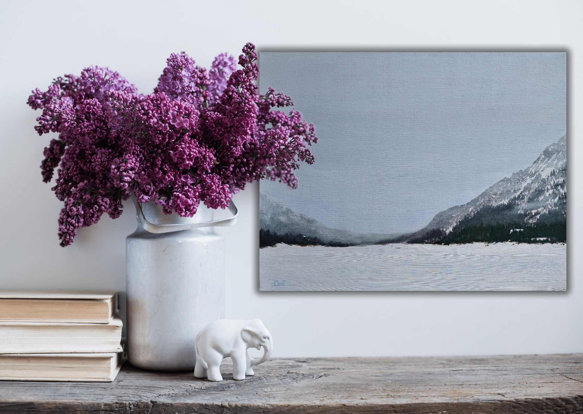 A wood table with books and purple lilacs in a pot with a landscape painting of mountains and a frozen lake in fog in the background
